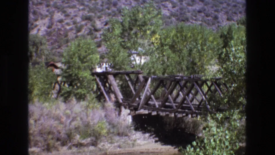 1973 New Mexico 8mm frame showing a rustic wooden truss bridge crossing a dry creek bed with trees and scrub below an arid hillside.