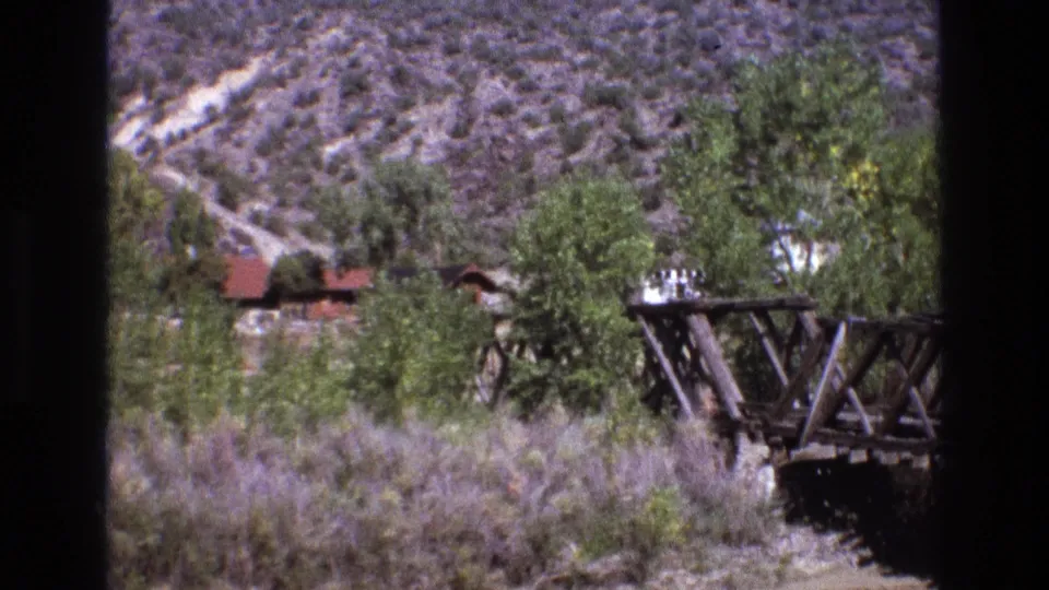 1973 New Mexico 8mm frame showing the wooden bridge at the edge of the shot, with scrub, trees, hillside, and a partially hidden red-roofed cabin-like structure behind it.