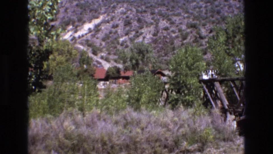 Final 1973 New Mexico frame with the bridge cropped at right, trees and brush across the foreground, a red-roofed rural structure in the distance, and an arid hillside behind.