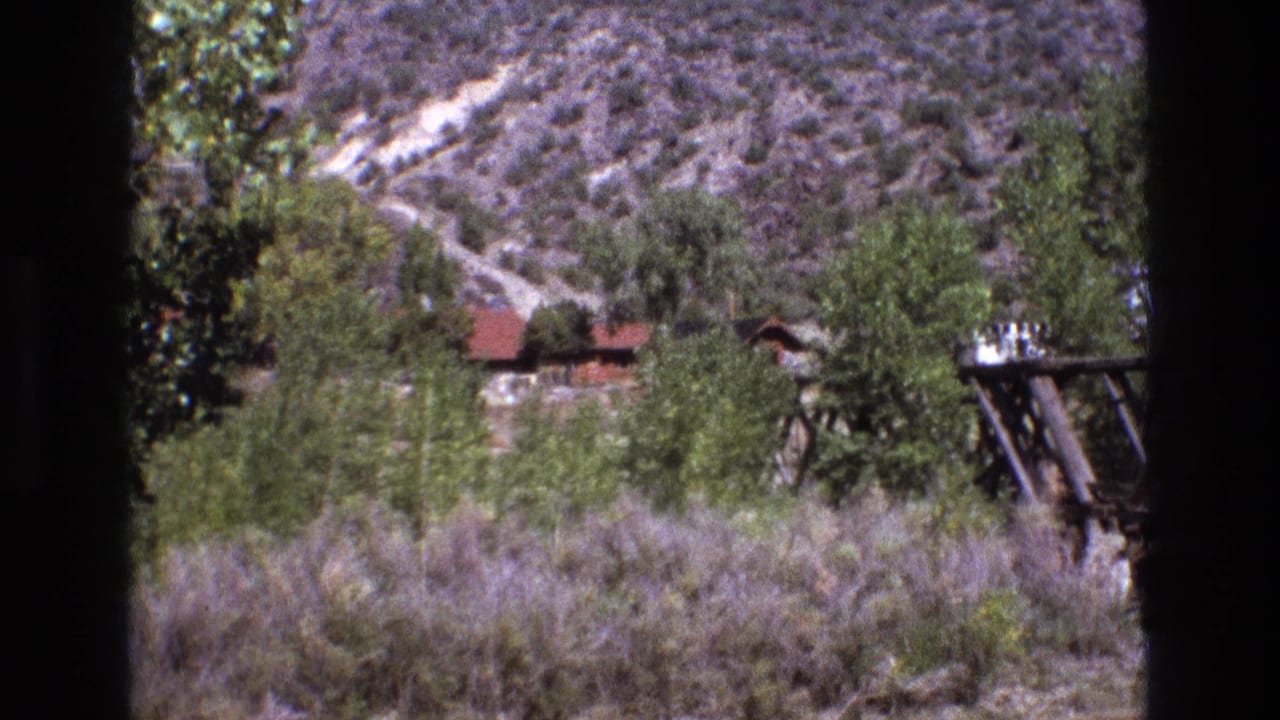 1973 New Mexico 8mm frame showing scrub-covered hills, green trees, a small red-roof cabin, and a wooden utility structure partly framed by dark edges.