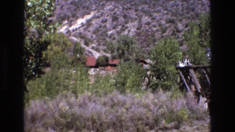 1973 New Mexico 8mm frame showing scrub-covered hills, green trees, a small red-roof cabin, and a wooden utility structure partly framed by dark edges.