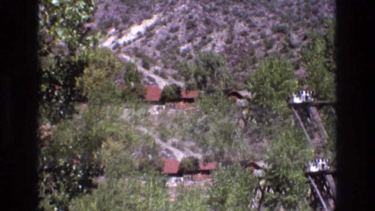 1973 New Mexico home-movie frame with dry hills, scattered greenery, red-roof structures, and a wooden structure at the right edge under clear sky.