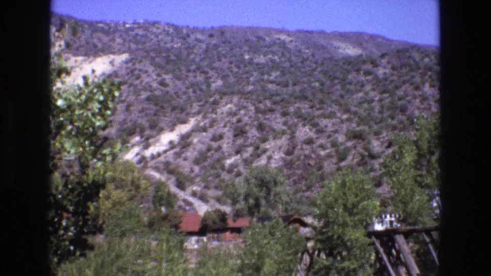 1973 New Mexico 8mm frame showing a wider scrub-covered hillside beneath blue sky, with trees and a small red-roof cabin near the lower edge.