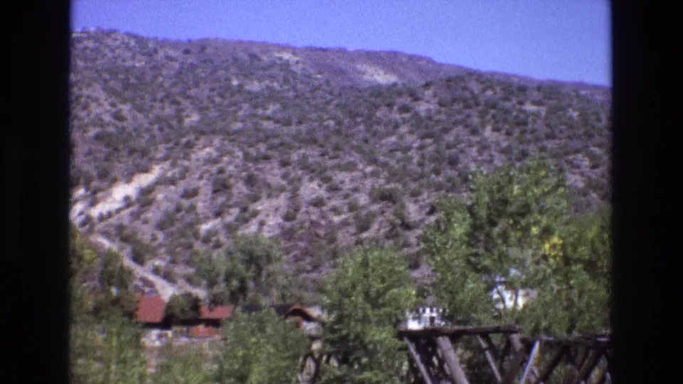 1973 New Mexico home-movie frame showing arid hills, a pale track, green trees, a red-roof cabin, and a partially visible wooden utility structure.