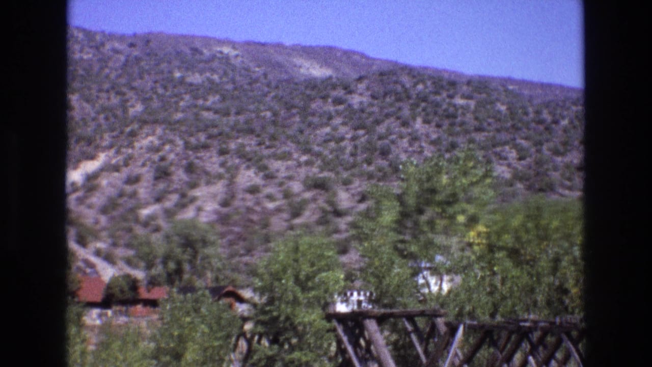 1973 New Mexico 8mm frame showing a scrub-covered hillside, scattered trees, and a weathered wooden bridge or trestle under bright daylight.