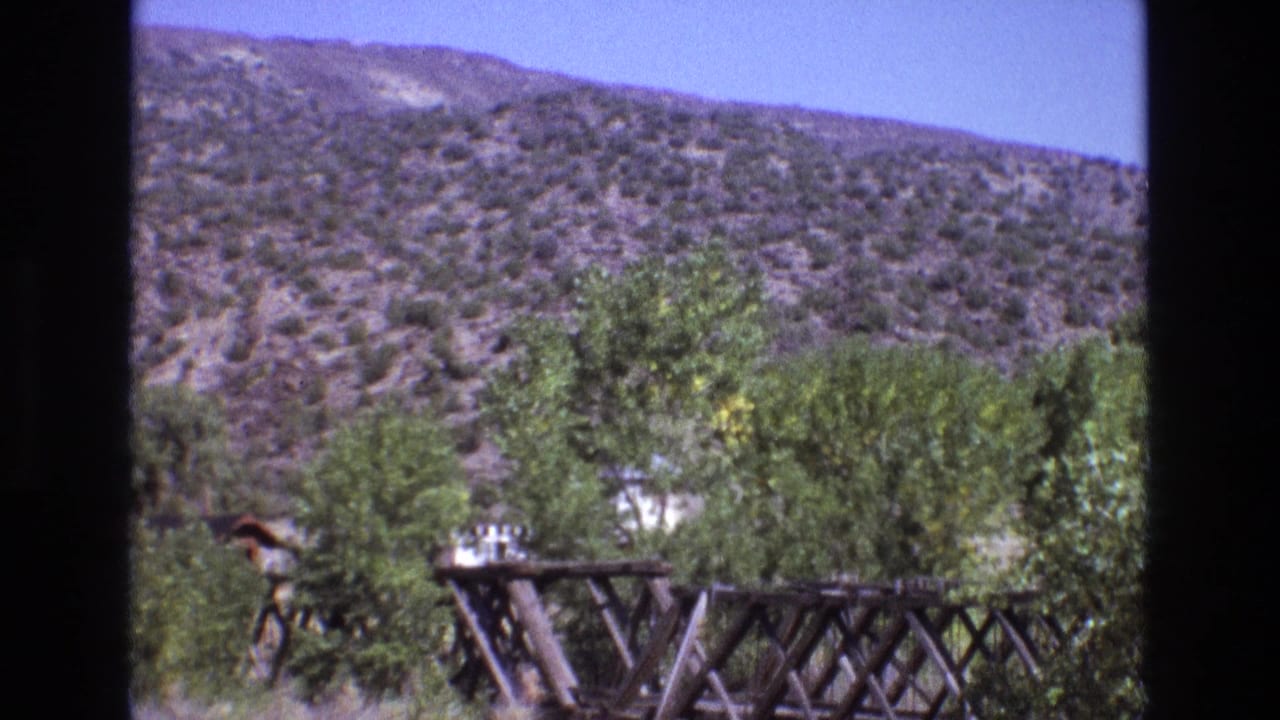 1973 New Mexico 8mm frame with a rustic bridge or trestle crossing the foreground, green trees beyond it, and dry brush-covered hills in the background.