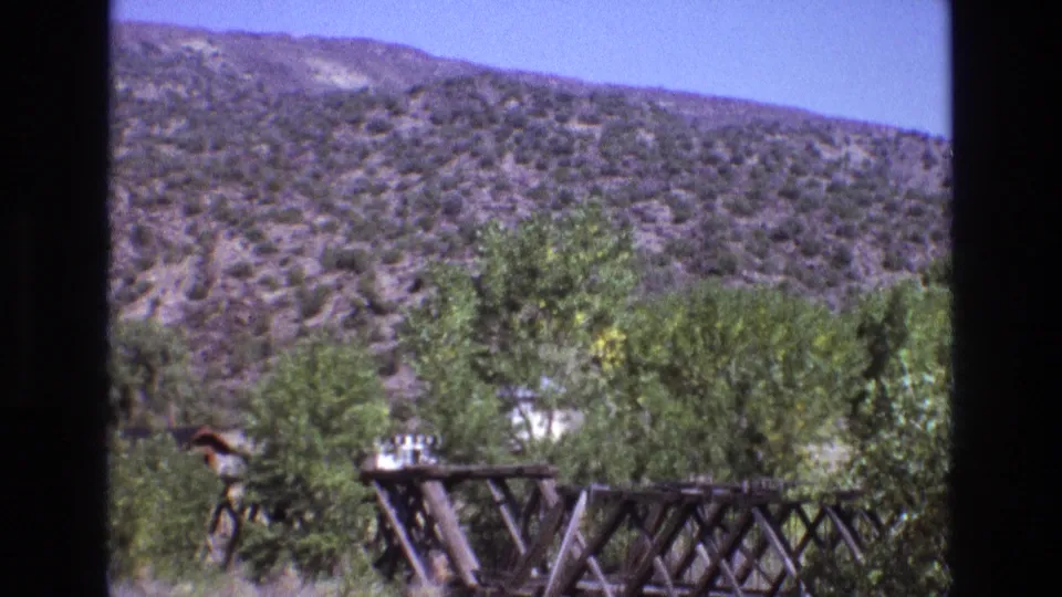 1973 New Mexico 8mm frame with a rustic bridge or trestle crossing the foreground, green trees beyond it, and dry brush-covered hills in the background.