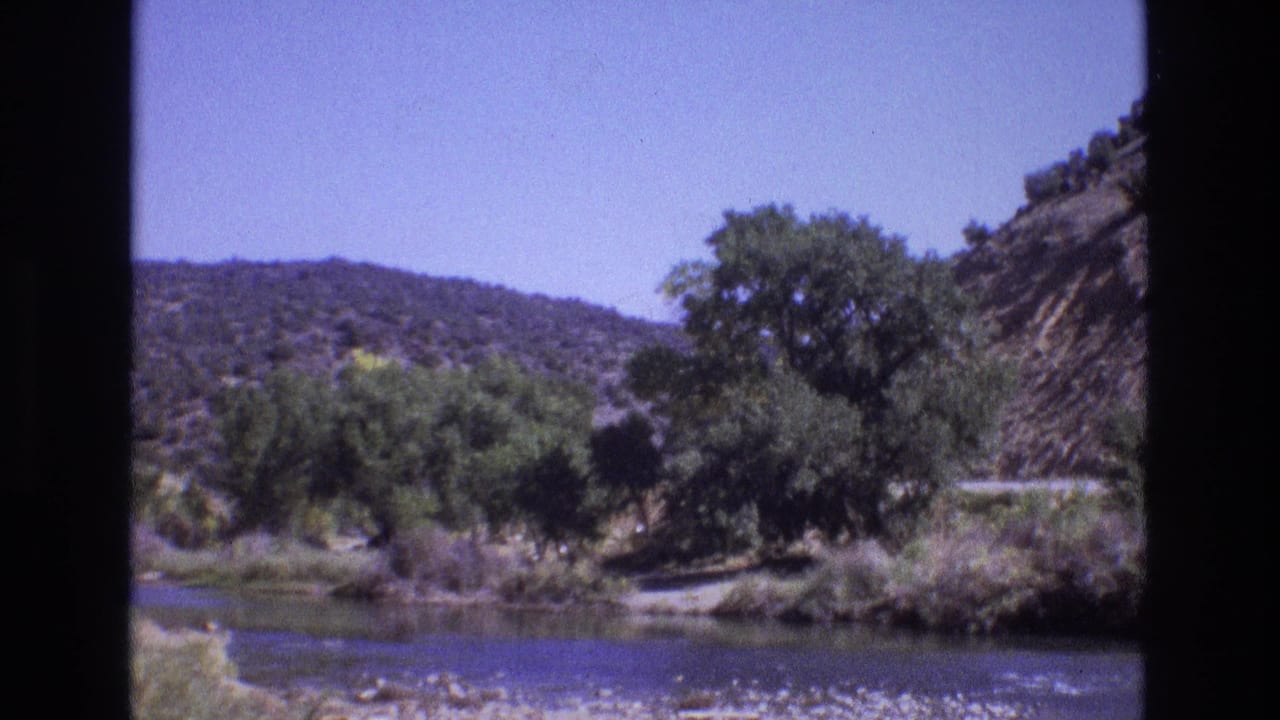 1973 New Mexico 8mm frame showing a quiet riverbank with green trees, dry hillside terrain, blue sky, and a road partly visible behind foliage.