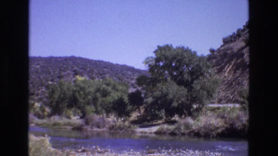 1973 New Mexico 8mm frame showing a quiet riverbank with green trees, dry hillside terrain, blue sky, and a road partly visible behind foliage.