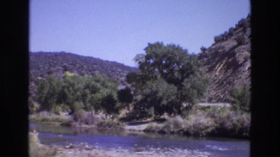1973 New Mexico 8mm frame showing a wider view of a river or stream bordered by green trees and scrub, with dry hills under clear blue sky.