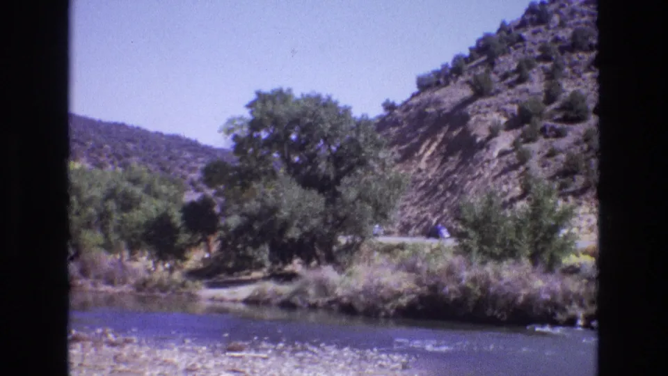 1973 New Mexico 8mm frame showing a river and roadside trees below a dry hillside, with a small vehicle briefly visible on the road at the right.