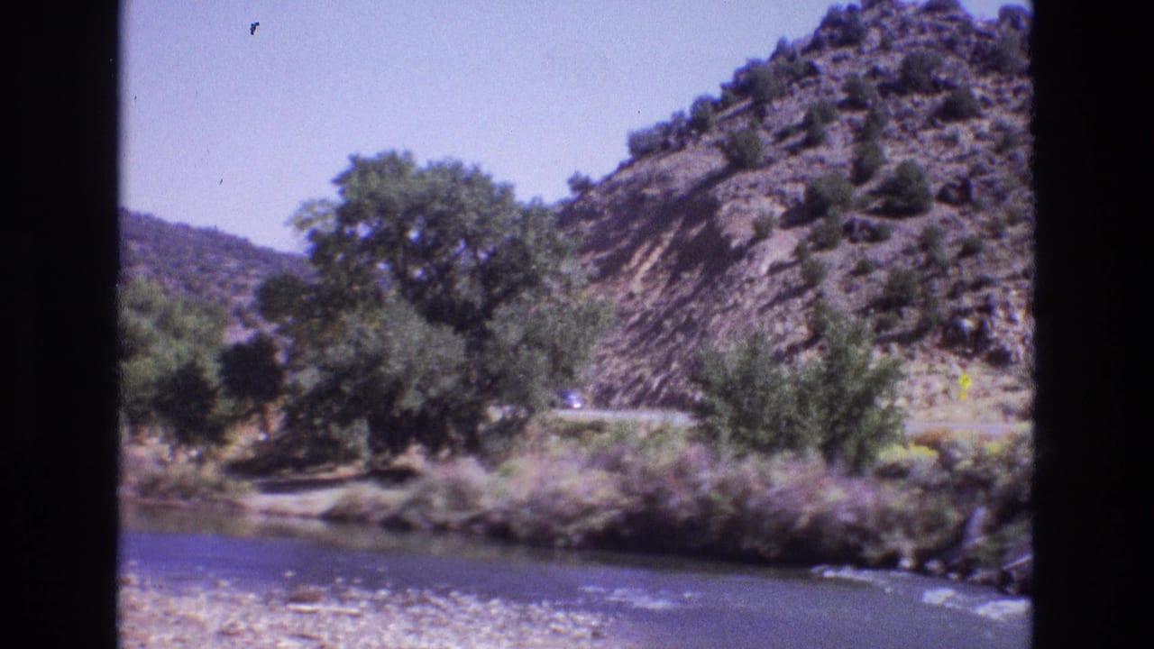 1973 New Mexico 8mm frame showing a river in the foreground, roadside trees, and a rocky hillside road seen through dark window or film borders.