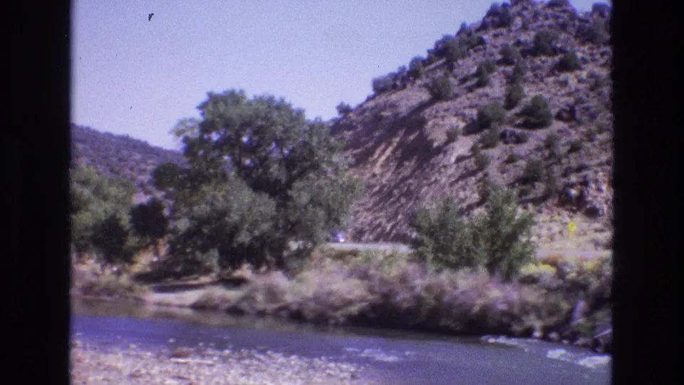 1973 New Mexico 8mm frame showing a river in the foreground, roadside trees, and a rocky hillside road seen through dark window or film borders.