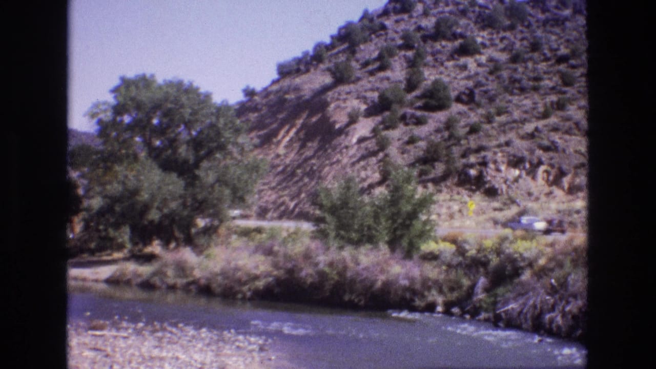 A car passes along a hillside road above a river and brushy bank.
