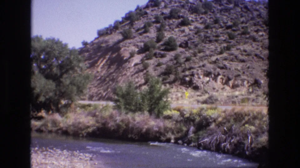 1973 New Mexico 8mm frame looking across a river toward roadside trees, scrub, and a rocky hillside beside a mountain road.