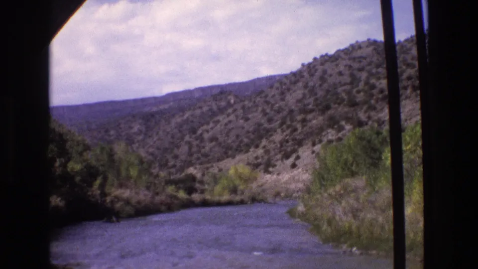 1973 New Mexico 8mm frame showing a river curving through scrub-covered hills, with trees along the bank and mountains in the distance.