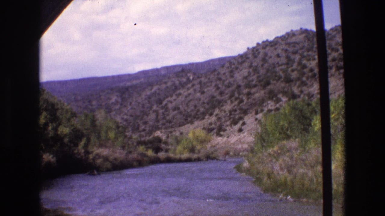 1973 New Mexico home-movie frame showing a wide river bordered by green trees and arid hills, viewed from inside a moving vehicle.