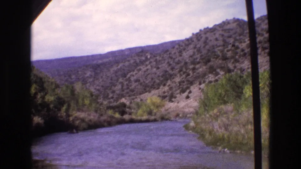 1973 New Mexico 8mm frame showing a river, roadside vegetation, and scrub-covered hills from a traveler’s window-framed viewpoint.