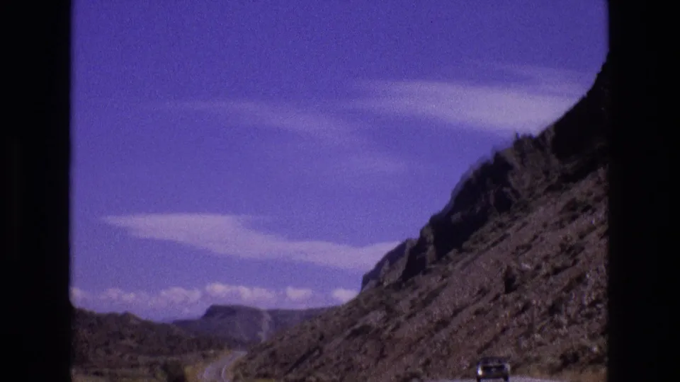 1973 New Mexico travel frame showing a road cut through steep rocky terrain, with a distant car and blue sky beyond the canyon-like slope.