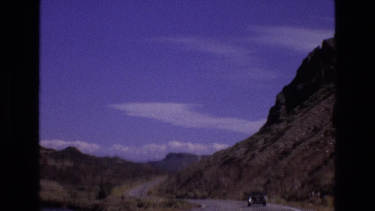 1973 New Mexico 8mm frame showing a winding road through rocky desert hills, sparse vegetation, and a dark vehicle in the distance.