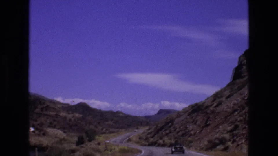 1973 New Mexico 8mm frame showing a road bending through rocky hills with a small dark car ahead and sparse roadside vegetation.