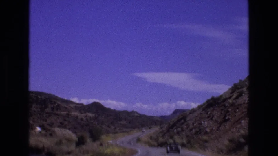 1973 New Mexico home-movie frame looking toward a curving road, rocky hillside, sparse vegetation, and a dark car near the lower right.