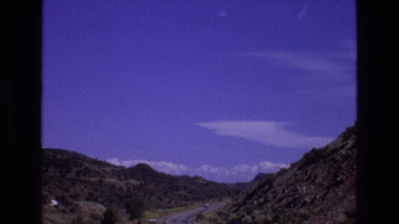 1973 New Mexico 8mm road-trip view of a paved road curving through arid hills, framed by dark window-like borders and a saturated blue-purple sky.