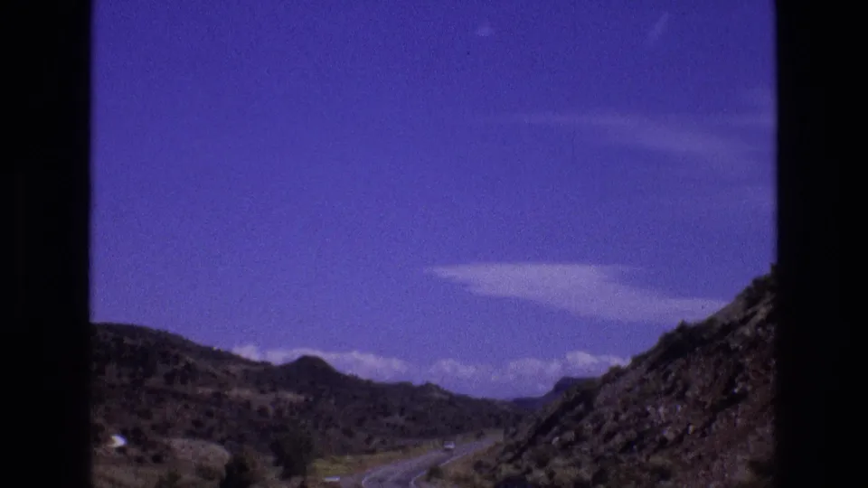 1973 New Mexico 8mm road-trip view of a paved road curving through arid hills, framed by dark window-like borders and a saturated blue-purple sky.