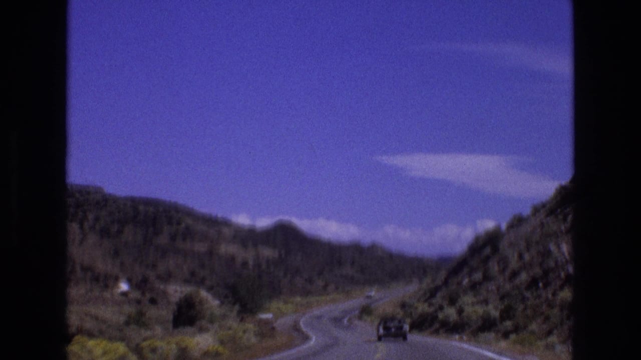 1973 New Mexico 8mm frame showing a distant vehicle on a winding mountain road between rocky arid hills beneath scattered white clouds.