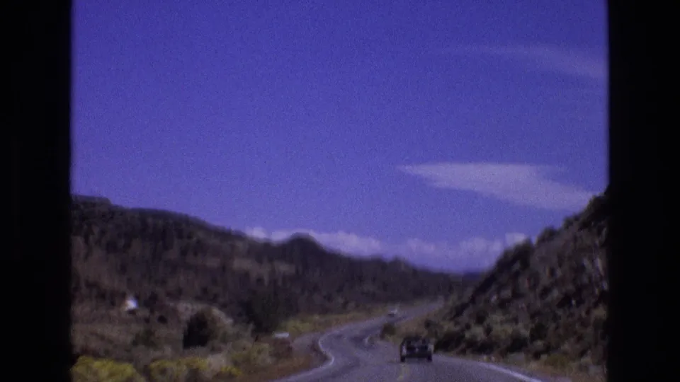 1973 New Mexico 8mm frame showing a distant vehicle on a winding mountain road between rocky arid hills beneath scattered white clouds.