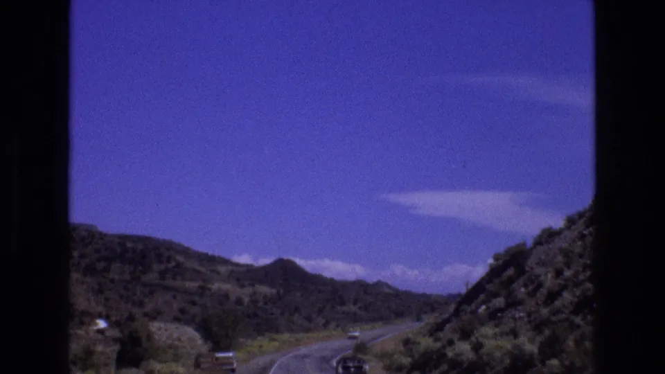 1973 New Mexico 8mm road scene with a vehicle on a curving paved route, low arid hills, sparse vegetation, and a saturated sky.