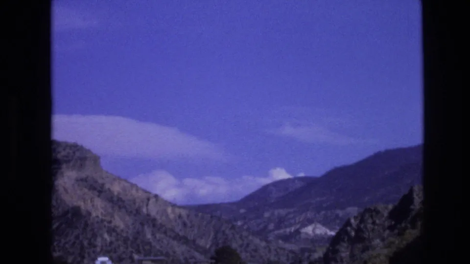 1973 New Mexico 8mm frame looking across a rugged mountain valley with arid slopes, a road edge, distant hills, and broad white clouds in a saturated sky.