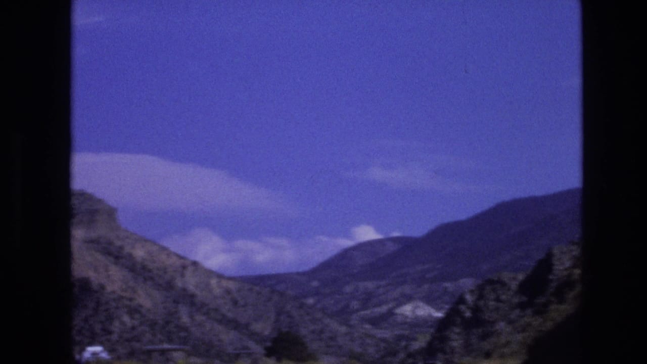 1973 8mm home-movie frame from New Mexico showing a mountain road landscape with rocky slopes, distant ridges, blue sky, and dark gate edges.