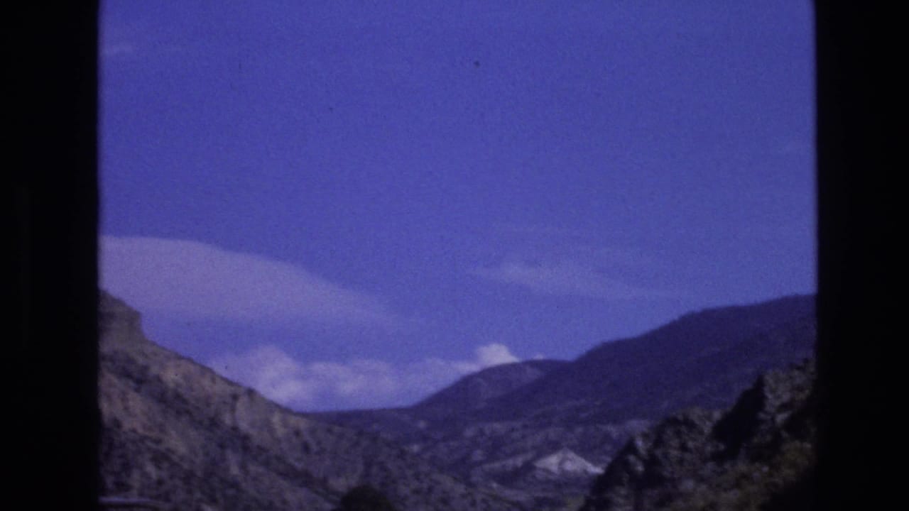 1973 New Mexico road-trip frame showing rugged hills and distant mountain ridges beneath a blue sky with scattered clouds.