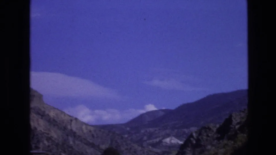 1973 New Mexico road-trip frame showing rugged hills and distant mountain ridges beneath a blue sky with scattered clouds.