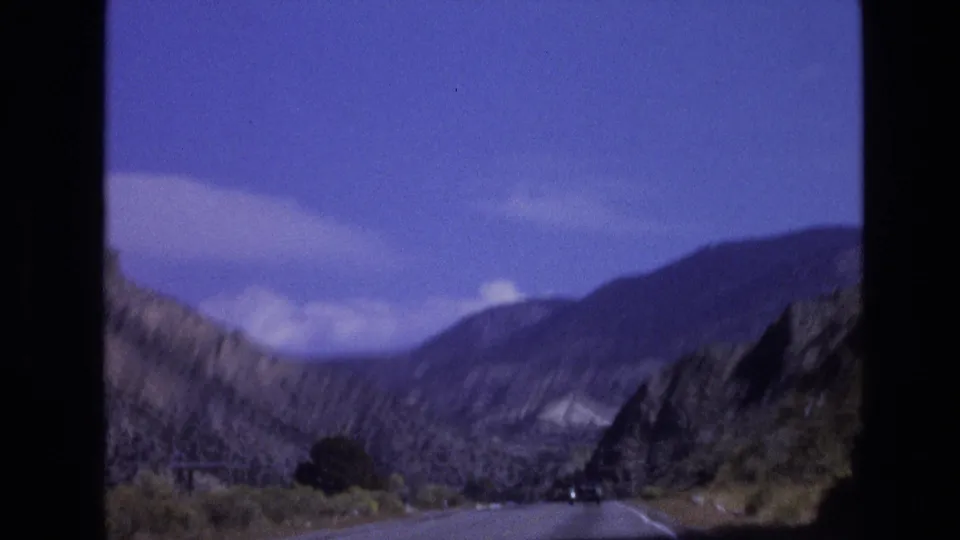 1973 8mm frame from a New Mexico mountain road trip, looking ahead toward a paved road, distant car, rocky hills, and blue sky.