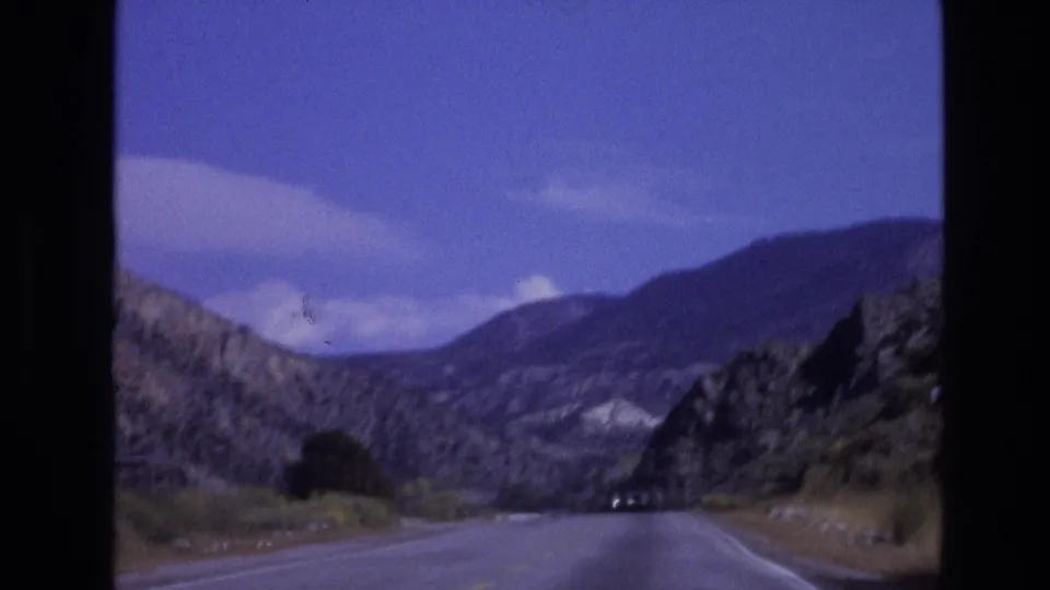 1973 New Mexico home-movie frame showing a paved road through arid rocky hills with distant mountains and scattered clouds overhead.