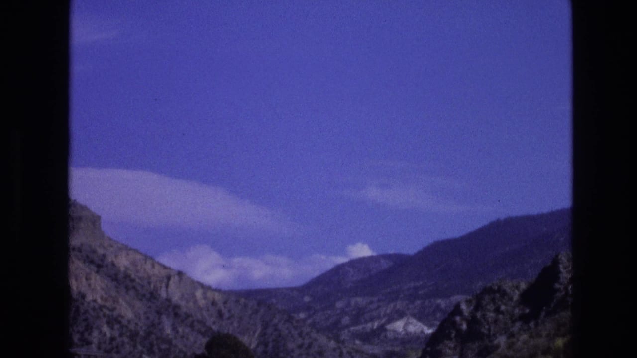1973 New Mexico 8mm frame showing rugged mountain ridges beneath a blue sky, partially framed by dark vehicle-window edges.