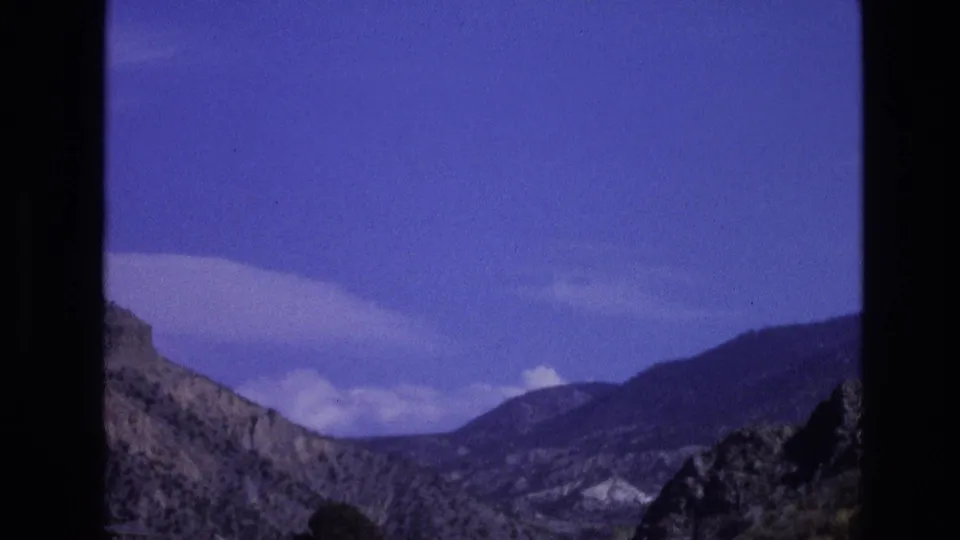 1973 New Mexico 8mm frame showing rugged mountain ridges beneath a blue sky, partially framed by dark vehicle-window edges.