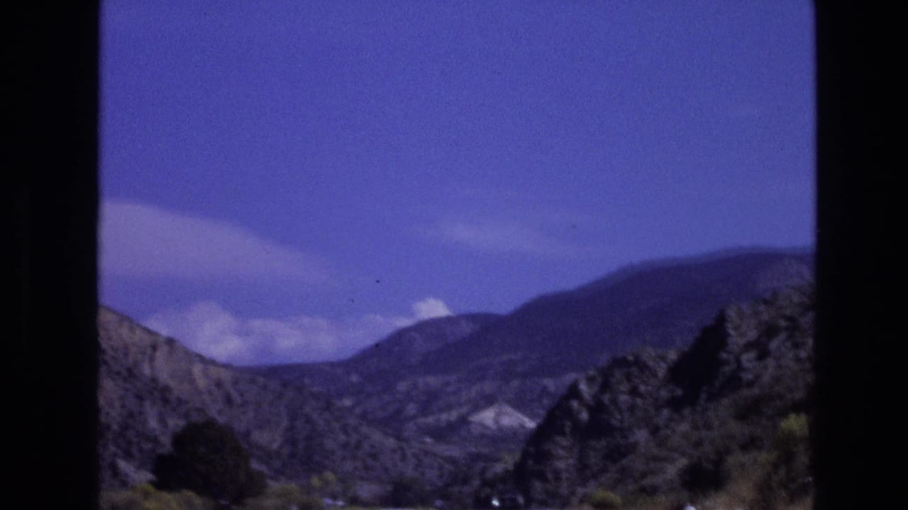 1973 New Mexico home-movie frame with distant hills, layered mountain slopes, and clouds seen from a vehicle window.