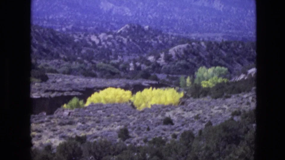 1973 New Mexico frame showing arid scrubland, a patch of bright yellow foliage, and layered mountain terrain in the distance.