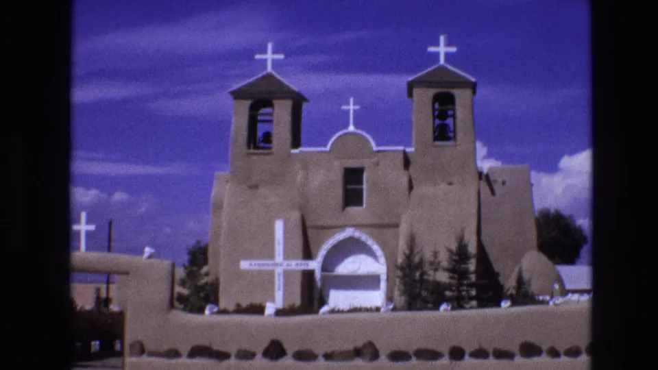 1973 New Mexico 8mm frame showing an adobe-style church exterior with twin bell towers, white crosses, a low wall, and vivid blue sky.