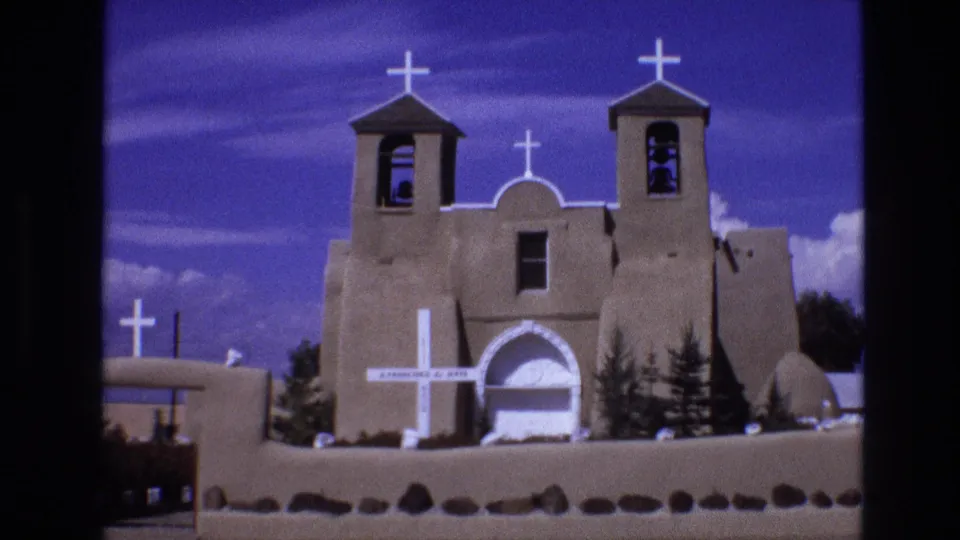 1973 New Mexico 8mm frame showing a closer visitor-shot view of an adobe-style church, twin bell towers, white crosses, arched doorway, and low stone wall.