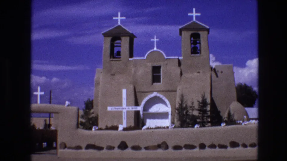 1973 New Mexico 8mm frame showing the adobe-style church exterior with twin towers, several white crosses, a low wall, small trees, and saturated blue sky.