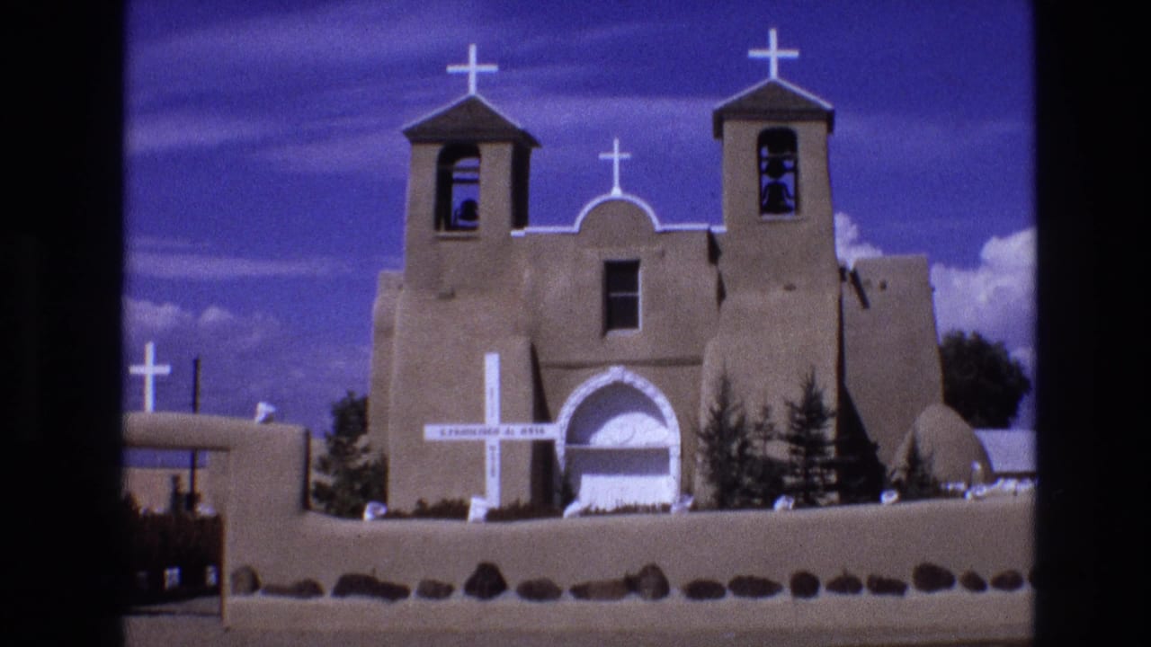 1973 New Mexico 8mm frame showing a tan Southwestern-style church with twin bell towers, white crosses, an arched entrance, and a low wall under a blue sky.