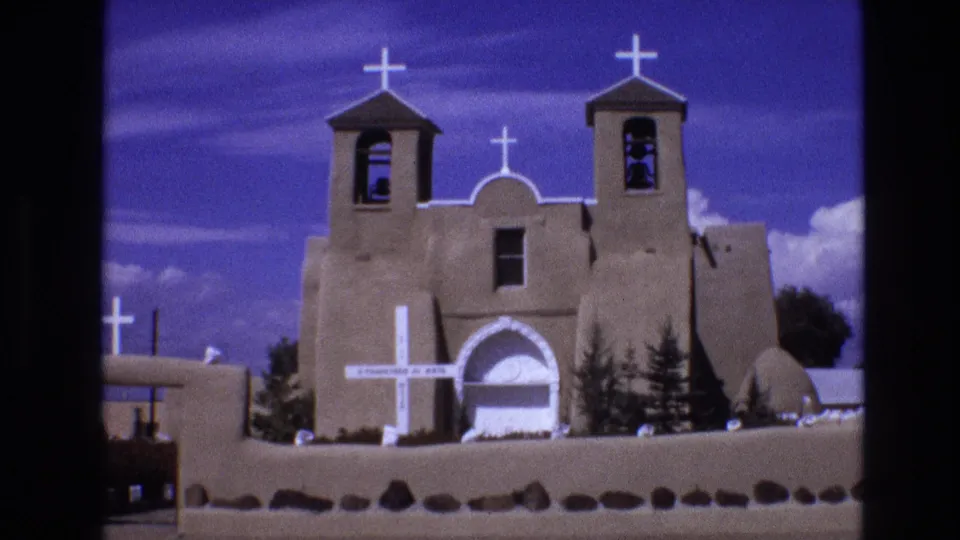 1973 New Mexico 8mm frame of a Southwestern-style church exterior with white crosses, two bell towers, a central arched doorway, and a low perimeter wall.