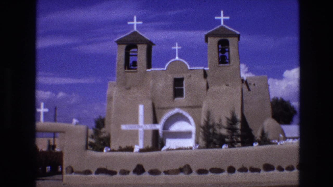 Adobe-style church facade with white crosses, twin towers, low wall, and foreground gate edge.