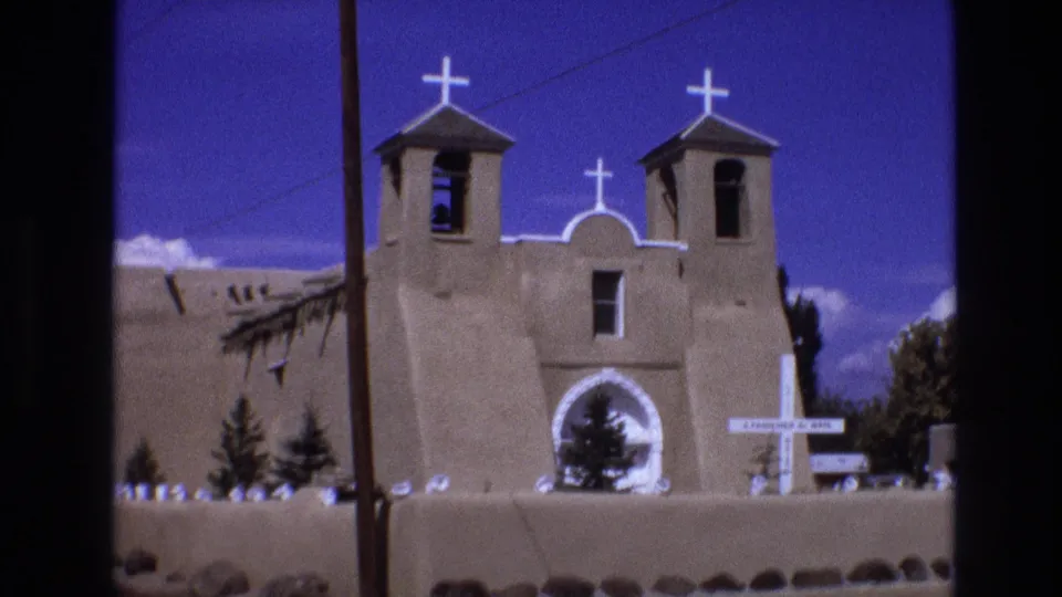 1973 New Mexico 8mm frame showing an angled view of the Southwestern-style church with white crosses, twin bell towers, a utility pole, low wall, and trees near the facade.