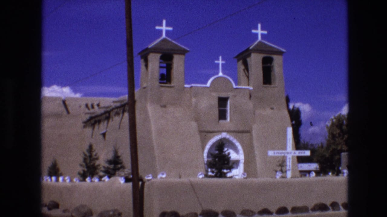 1973 New Mexico 8mm frame showing an adobe church facade with twin bell towers, white crosses, a central arched entrance, a low wall, small trees, and a utility pole at the left.