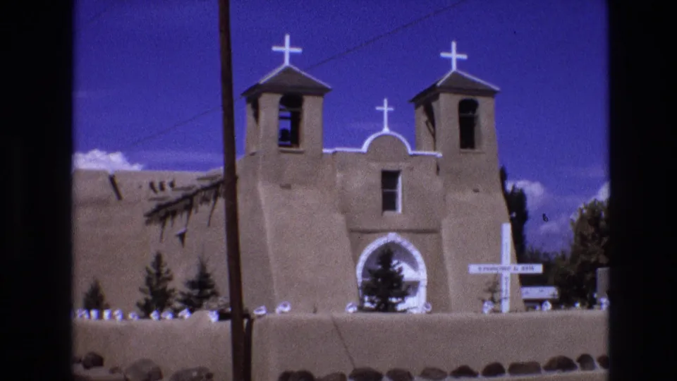 1973 New Mexico 8mm frame showing an adobe church facade with twin bell towers, white crosses, a central arched entrance, a low wall, small trees, and a utility pole at the left.
